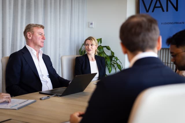 In a boardroom, Vantage Asset Management colleagues discuss around a table with a laptop, plants, and a company poster visible.