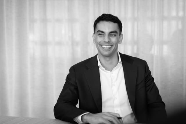 A man in a dark suit smiles while seated at a table against a sheer curtain, in this black and white portrait for Vantage Asset Management.