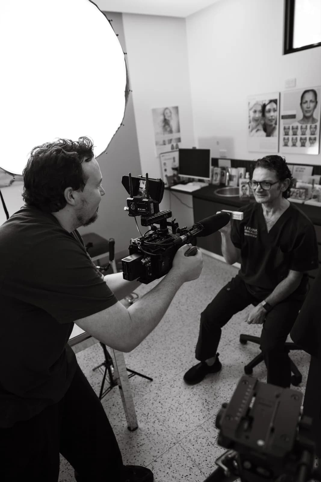Camera operator filming a seated subject during a talking-head interview with a softbox lighting the room.
