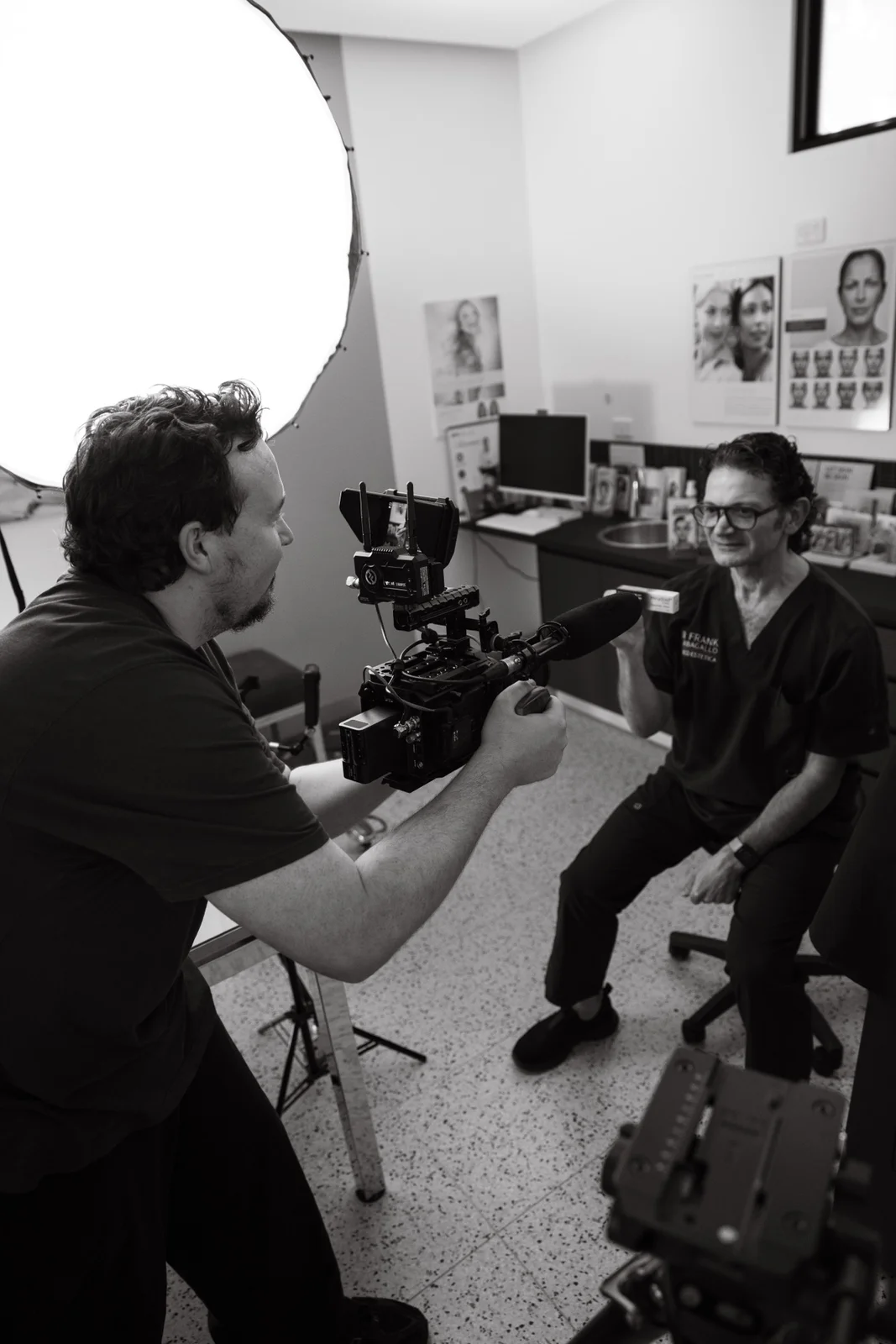 Camera operator filming a seated subject during a talking-head interview with a softbox lighting the room.